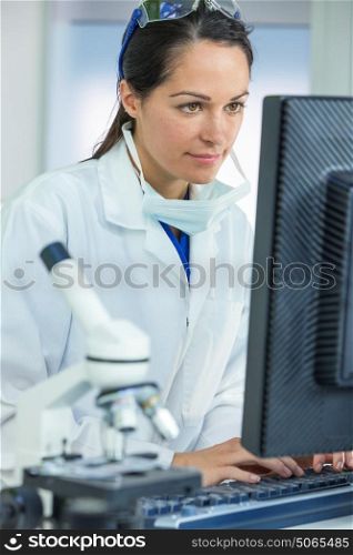 A thoughtful female medical or scientific researcher or woman doctor using a computer in a laboratory with microscope and other equipment in the foreground.