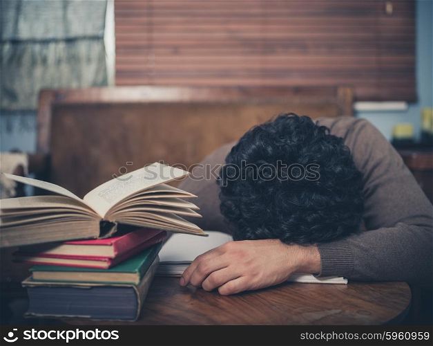 A tired student is asleep with his head on a coffee table surrounded by books