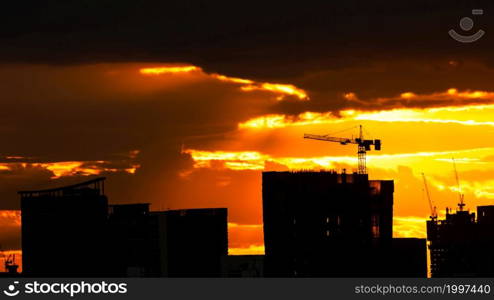 A tower crane lifting on the roof of building at a construction site. Dramatic strom cloud on the sunset sky in the backgrounds. The skyscrapers under construction. Bangkok, Thailand.