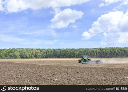 A tractor in a field shallowly plows the soil with metal discs after harvesting against a blue cloudy sky.. Tractor in the field cultivates the soil after the harvest.