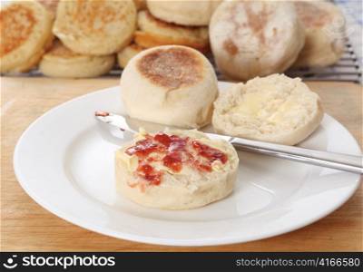 A traditional English muffin tea-bread with butter and jam in front of a tray of fresh-baked home-made muffins and crumpets