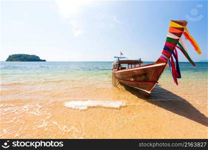 A traditional wooden boat on a beach on sunny summer, traditional colors cloth hanging on the bow of the boat, an island in the background. South Thailand.