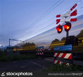 A train passing a rail crossing, surrounded by fences and barb wire on a summer evening.