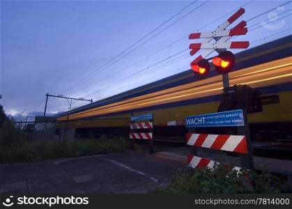 A train racing past a pedestrian crossing just after dusk