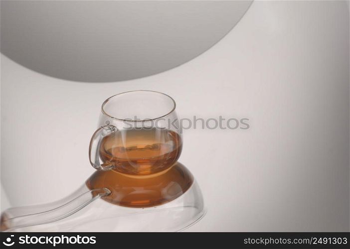 A transparent glass cup with tea on a reflecting surface and a light gray background. Transparent glass teapot and cup with tea