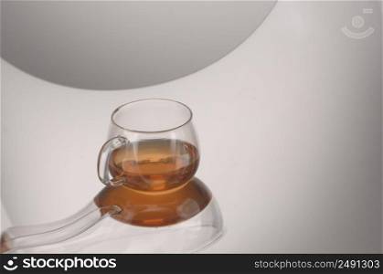 A transparent glass cup with tea on a reflecting surface and a light gray background. Transparent glass teapot and cup with tea
