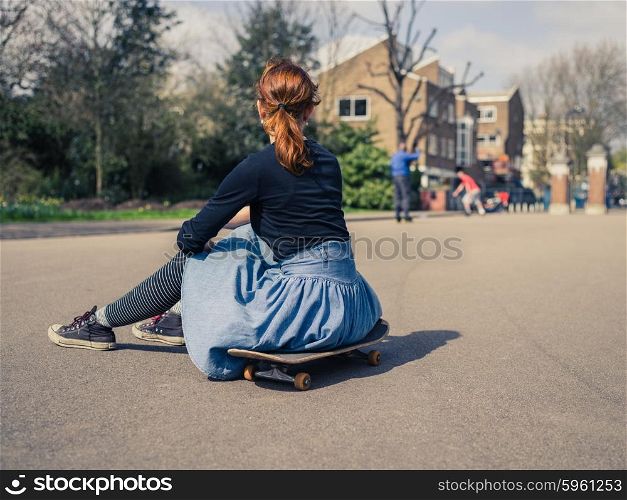 A trendy young woman is sitting on a skateboard in a park and is watching other people skateboard in the distance