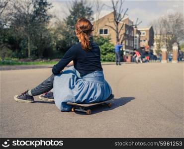 A trendy young woman is sitting on a skateboard in a park and is watching other people skateboard in the distance
