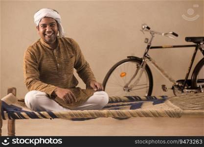 A TURBANED VILLAGER HAPPILY SITTING ON A COT AND LOOKING AT CAMERA