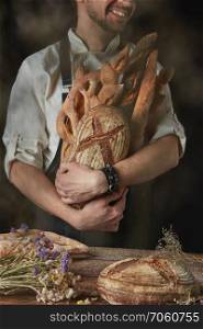 A variety of rustic organic bread in the hands of a man on a dark background and old wooden table with dried flowers and bread. Baker keeps a variety of bread