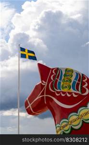 A vertical view of a colorful Swedish Dala horse and the Swedish flag under an expressive sky