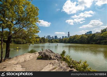 A view of a lake in Central Park, New York City, on a summer day, with people enjoying it. On the background, buildings and skyscrapers can be seen.