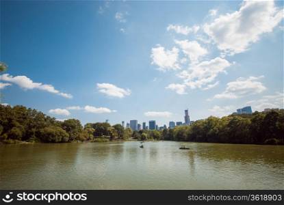 A view of a lake in Central Park, New York City, on a summer day, with people enjoying it. On the background, buildings and skyscrapers can be seen, which are also reflected on the water.