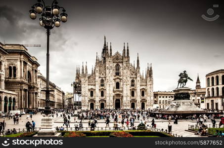 A view of Milan Cathedral fromthe front, including the large square filled with people in front of it, and some buildings to the side.