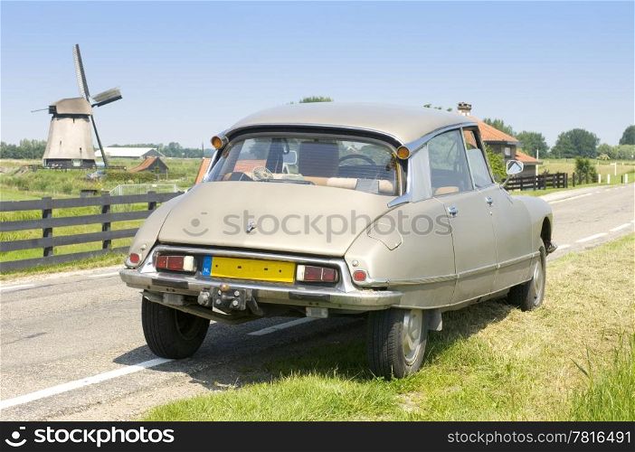 A vintage 1973 Citroen DS parked on a country road with a windmill in the distance on a bright summer day.