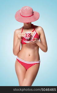 A vintage photo of a beautiful pin-up girl in bikini drinking tea and hiding head under a sun hat.