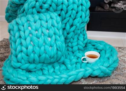 A warm plaid knitted from merino wool of a beautiful aquamarine shade lies near the fireplace along with a cup of tea. The concept of comfort. A warm plaid knitted from merino wool of a beautiful aquamarine shade lies near the fireplace along with a cup of tea. The concept of comfort.