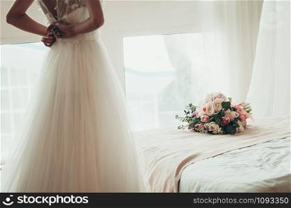 A wedding bouquet on a white bed, with blurred bride buttoning her dress, a view from her back