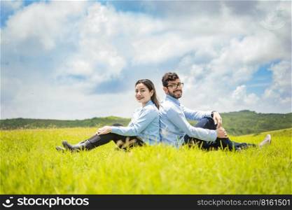 A wedding couple sitting with their backs to each other on the grass, Wedding couple in the field sitting with their backs to each other looking towards the camera