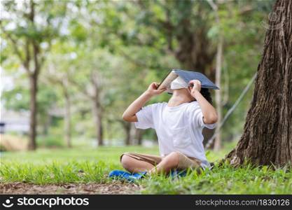 A white boy wearing a hygienic mask sits alone under a tree reading a book covering his head.