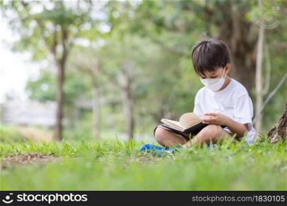 A white boy wearing a hygienic mask sits alone under a tree reading a book.