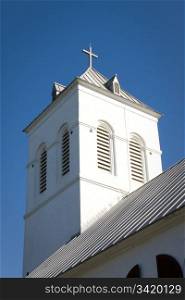 A white cross sits on the top of a church bell tower against a blue sky.