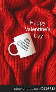 A white cup with a heart stands on a red knitted fabric. Inscription of St. Valentine&rsquo;s day, 14 February. A white cup with a heart stands on a red knitted fabric. Inscription of St. Valentine&rsquo;s day, 14 February.