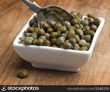 A White Dish Full Of Capers, With A Spoon Being Put In To Them, Up On A Wooden Surface