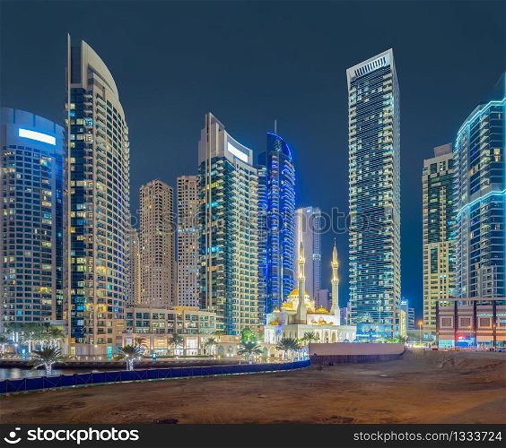 A white mosque near Dubai marina in downtown skyline, United Arab Emirates or UAE. Financial district and business area in smart urban city. Skyscraper and high-rise buildings at night.