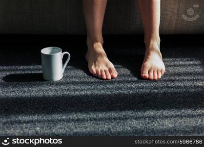 A white mug on the carpet next to a woman&rsquo;s feet as she is sitting on a sofa
