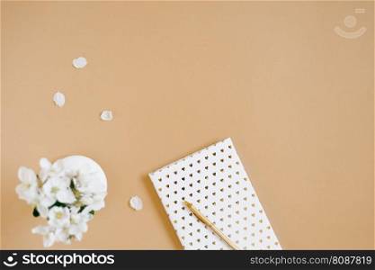 A white notebook with a gold pen on a beige table and flowers in a defocus vase. Blogger’s workplace. Top view, copy space