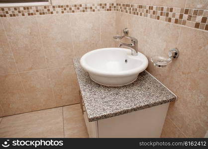 A white round basin positioned on a white granite topped build in cupboard inside a bathroom that is tiled with light brown ceramic tiles from top to bottom in a brand new home.