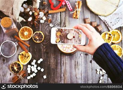 a white smartphone in a woman&rsquo;s hand takes a photograph of a mug with a drink and pieces of marshmallow on a gray wooden background, the top view