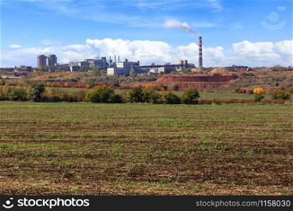 A wide agricultural field after harvesting against the background of a cement plant industrial complex with a quarry on a bright autumn day against the background of a slightly cloudy blue sky.. Agricultural field against the background of a cement plant and a quarry of limestone with clay on a summer day.
