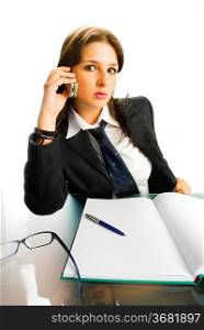 a wide view of a young woman in black jacket and white shirt and a tie sitting down behind an office table and talking by phone
