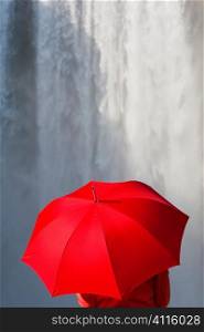 A woman dressed in a red jacket and carrying a red ubmrella standing in front of a waterfall. Shot on location at Skogafoss in Iceland.