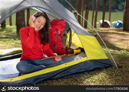 A woman Enjoy listening to music from headphones with play tablet. She sat in a yellow tent. She backpacking camping In the middle of a pine forest beside Lake. Pang Oung, Mae Hong Son, Thailand. Women listening to music camping tent.