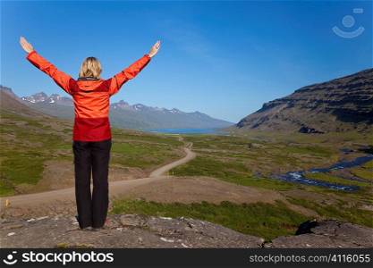 A woman hiker arms raissed in celebration looking down the Berufjordur Valley in Iceland. The Berufjor?ur fjord is visible at the end of the valley.