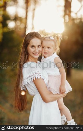 a woman holds a child in her arms in the park. young blonde mother holding her toddler daughter in her arms as they are walking through the park on a sunny day. little girl with two tails. a woman holds a child in her arms in the park.