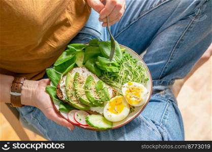 A woman holds a plate with useful healthy foods avocado, cucumber, spinach, radish, egg, microgreens, seeds. Proper healthy eating, keto diet. Green vegan breakfast. Close-up selective focus. A woman holds a plate with useful healthy foods avocado, cucumber, spinach, radish, egg, microgreens