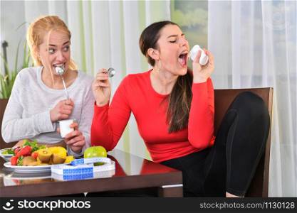 A woman hungrily finishes her yogurt while the other looks at her in amazement, sitting at a table with assorted fruit and a measuring tape. Healthy lifestyle concept.. Women finishing their yogurt and having fun