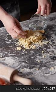 A woman in a black apron kneads the dough by adding water to the kitchen table. Preparation of dough for baking. On a kitchen table the women&rsquo;s hands kneads the dough. Step-by-step preparation of the dough