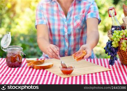 A woman in a plaid shirt smears jam on bread with a spoon. Red tablecloth, picnic basket, grapes. Sunny day. Picnic concept. A woman in a plaid shirt smears jam on bread with a spoon