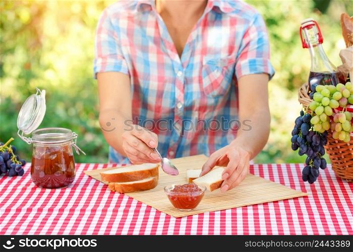 A woman in a plaid shirt spreads jam on bread. Red tablecloth, picnic basket, grapes. Sunny day. Picnic concept. A woman in a plaid shirt spreads jam on bread