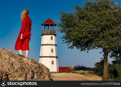 A woman in a red dress is standing on the beach near the lighthouse at sunrise.. A woman in a red dress is standing on the beach near the lighthouse at sunrise