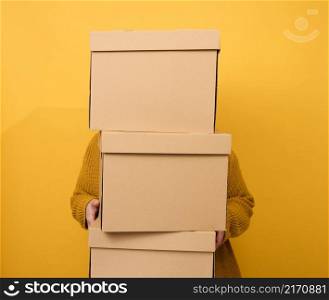 a woman in a sweater holds a stack of brown cardboard boxes on a yellow background. Relocation