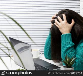 a woman in a sweater sits at the table with her head clasped in her hands, stress and headache from overwork, fatigue. A laptop and a cup of coffee are on the table