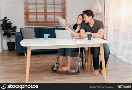 A woman in a wheelchair after a car accident and working with a computer at home with her lover. The concept of Mutual care and new technology has made people with disabilities Equality in society.