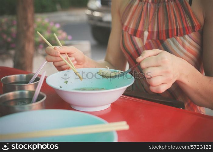A woman is sitting at a table in the street and is eating noodles