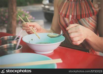 A woman is sitting at a table in the street and is eating noodles